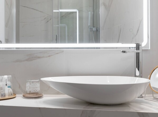 Un baño moderno con un lavamanos grande de color blanco, cabina de ducha con vidrio y pared con azulejos de mármol.