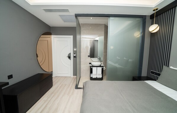A modern hotel room with a bed, a bathroom with a sink and mirror, a walk-in closet, and a sliding door to the bathroom.