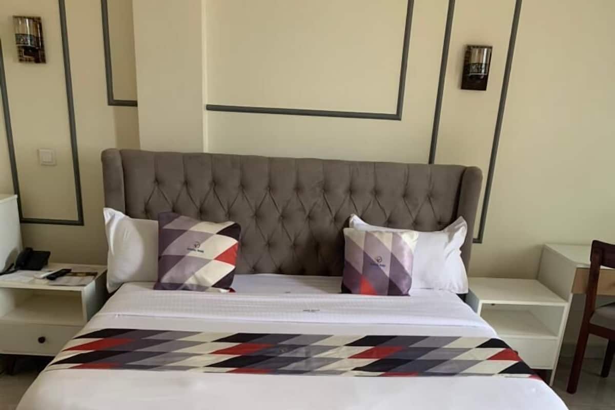 Una cama con cabecera capitonada, dos almohadas, una colcha y una mesita de noche con una lámpara.