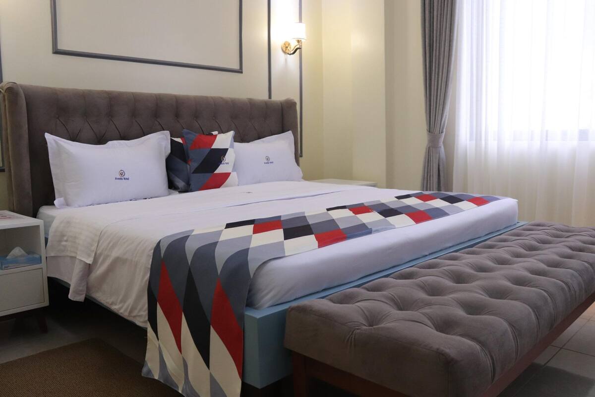 Una cama bien hecha con colcha a cuadros, un banco tapizado, una mesita de noche con lámpara y un cuadro en la pared.