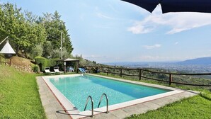 Ein rechteckiger Swimmingpool mit klarem, blauem Wasser, umgeben von einer Steindeck und Rasenflächen. Liegestühle und ein kleines Pavillon sind vorhanden. Ein Holzzaun schließt das Areal ein, und im Hintergrund sind Hügel und eine Stadtlandschaft zu sehen.