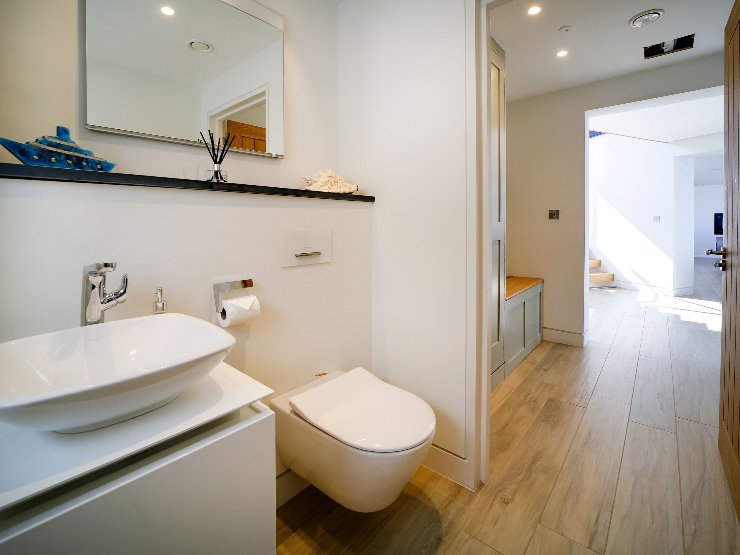 Ein modernes Badezimmer mit Waschbecken, Toilette und Spiegel in Weiß. Ein Holzboden und eine Tür zu einem anderen Raum sind ebenfalls vorhanden.
