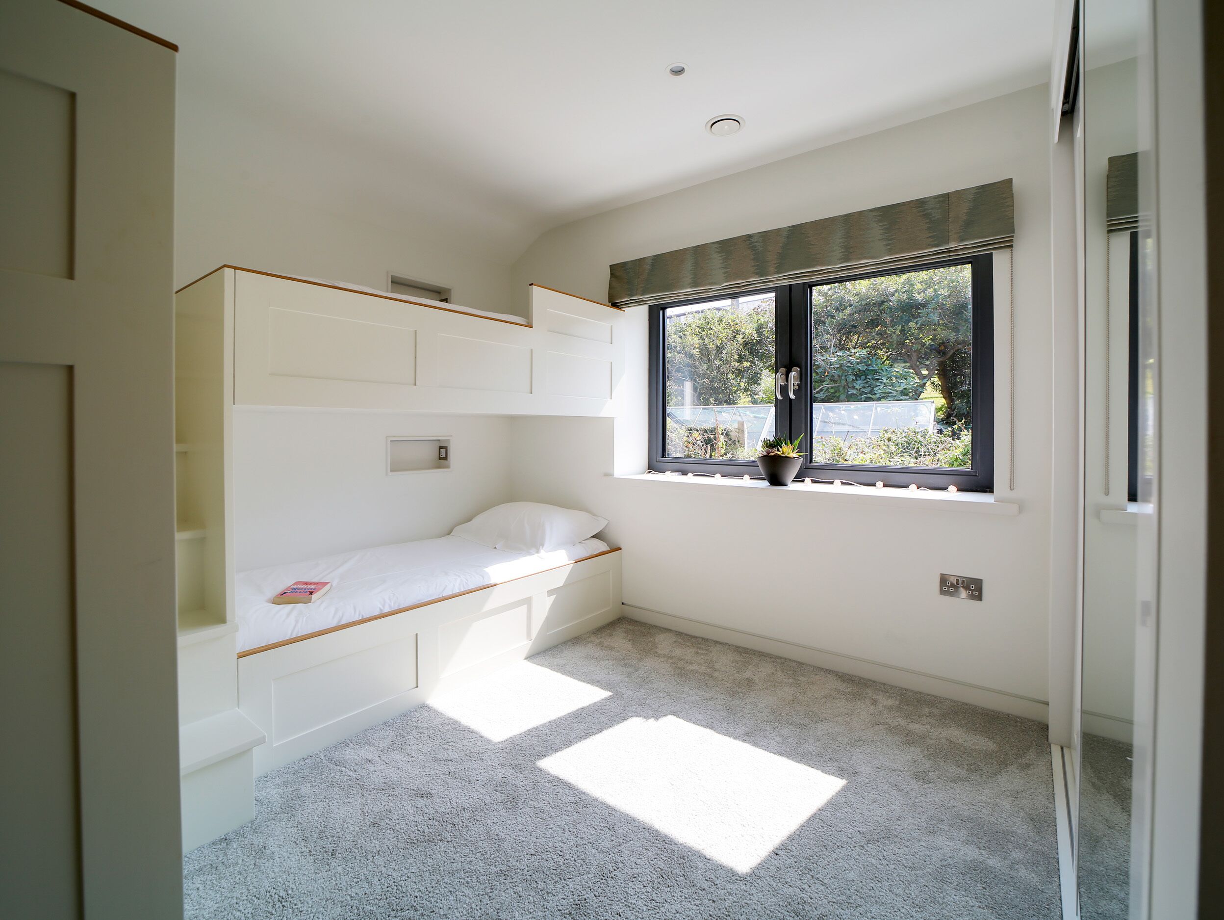 A small, bright room with a single bed, built-in storage, a window with a view, and a carpeted floor.