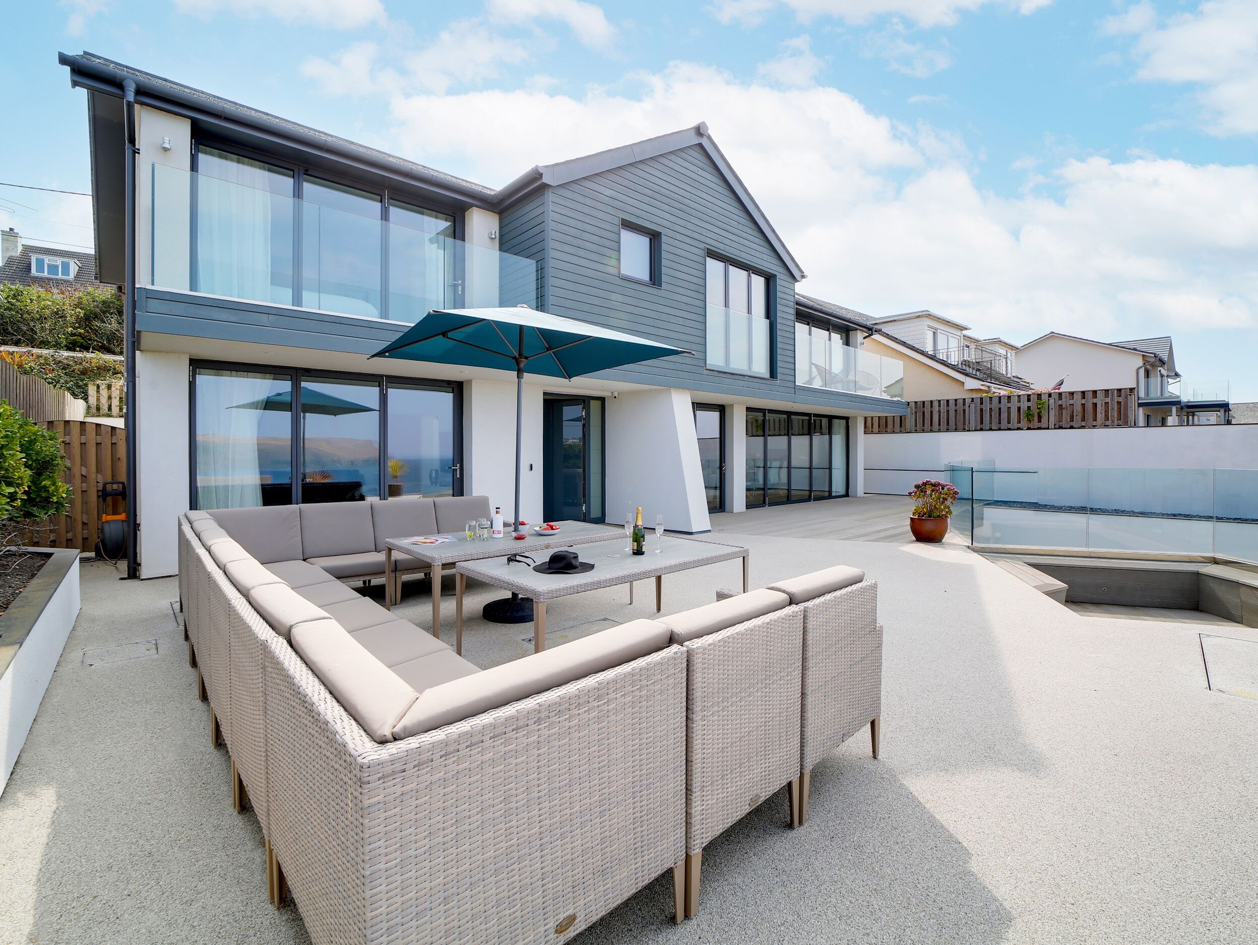 Une maison moderne dotée d’un grand patio comprenant un canapé en osier, une table à manger et un parasol bleu.