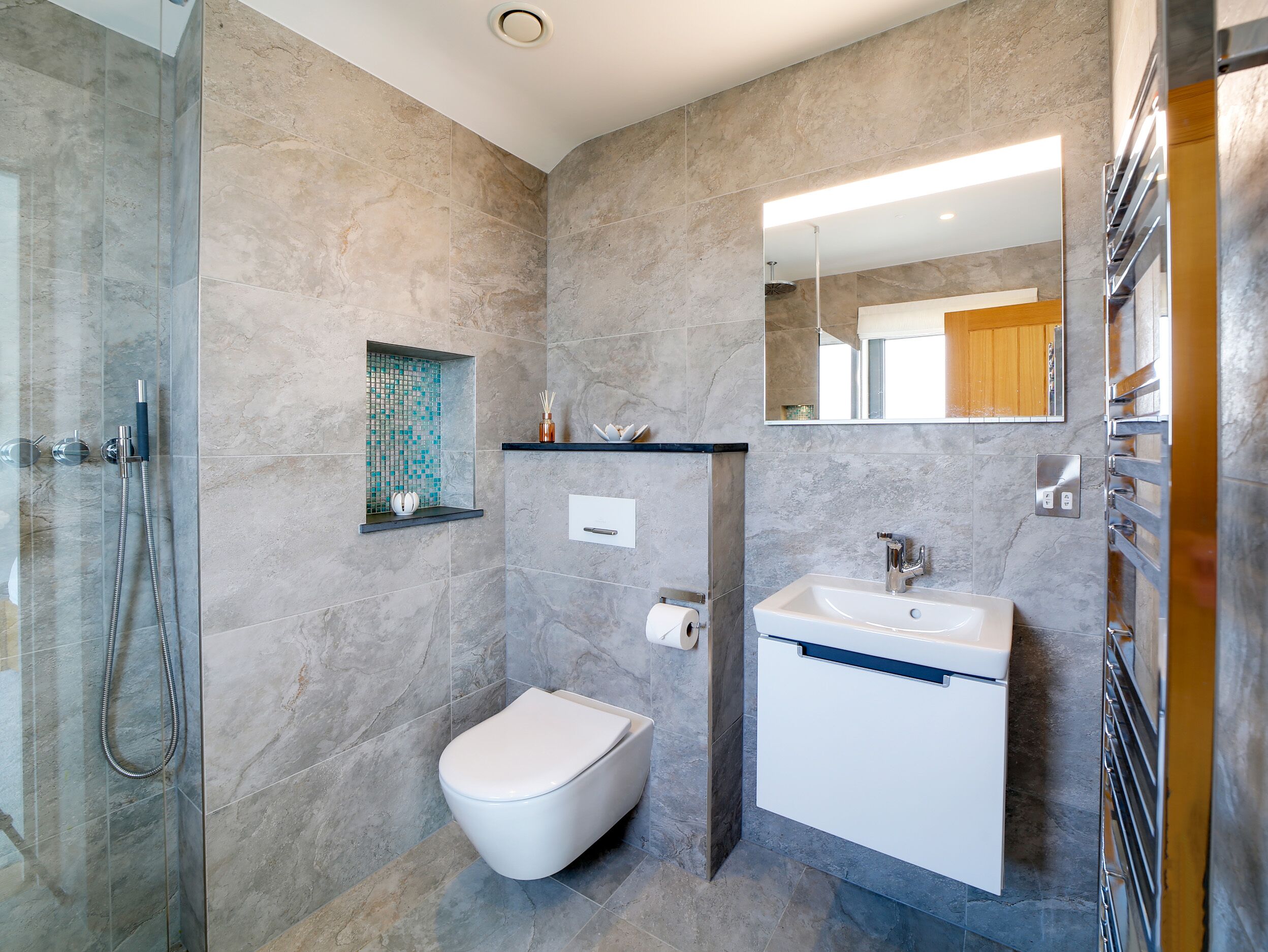 Une salle de bain moderne avec une douche à parois de verre, des toilettes blanches, un lavabo avec un miroir au-dessus et une petite étagère contenant des articles de toilette.