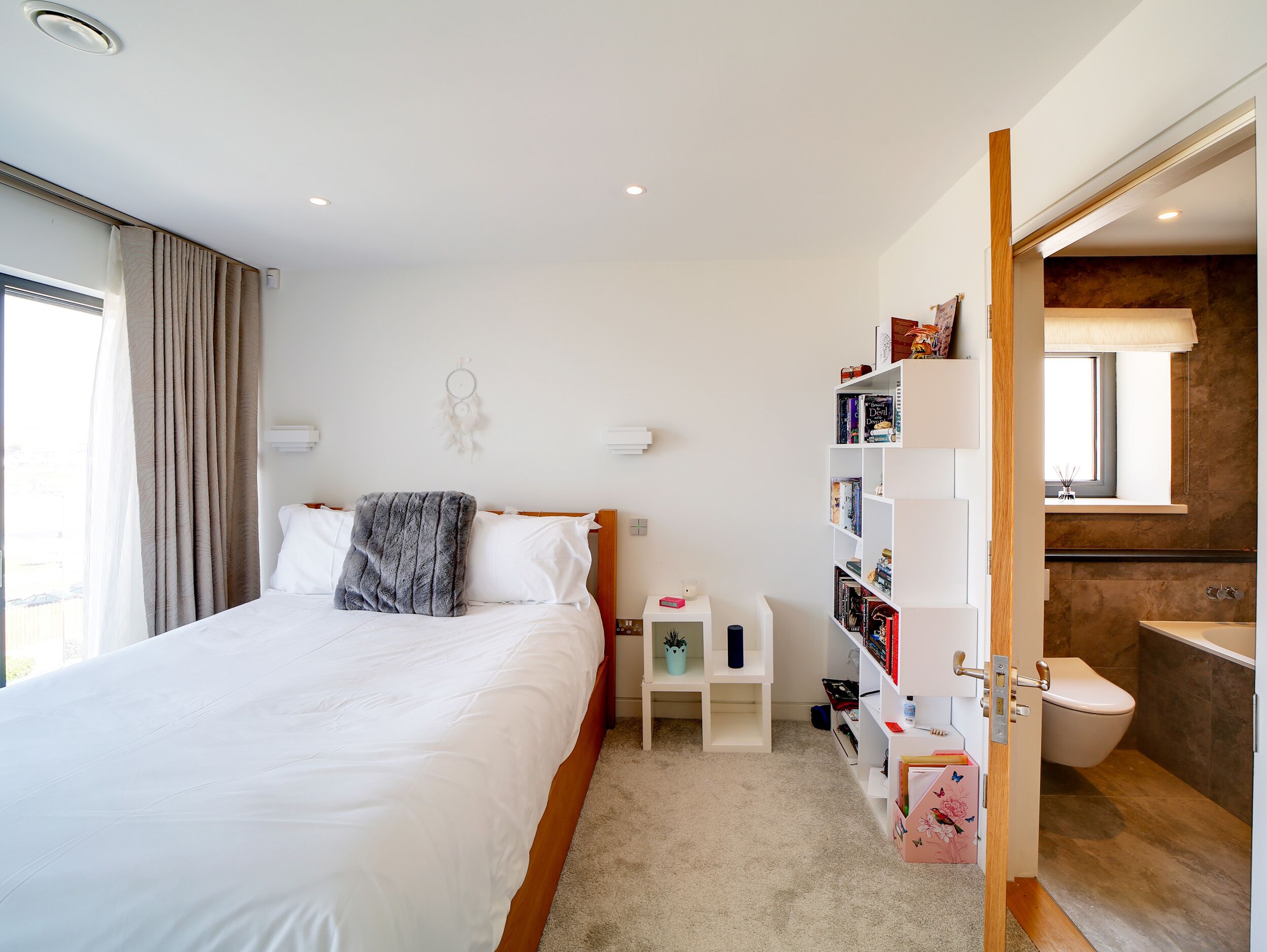 A bedroom with a large bed, a bedside table, a bookshelf, and a bathroom with a toilet and sink.