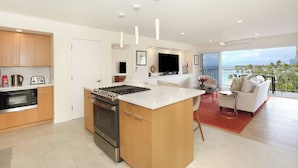 A modern kitchen with a central island, built-in oven, and microwave. There is a dining area with a sofa and a TV. The room has large windows offering a view of the city.