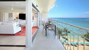 A balcony with a view of the ocean, a dining area with chairs and a table, and a living room with a sofa and TV.