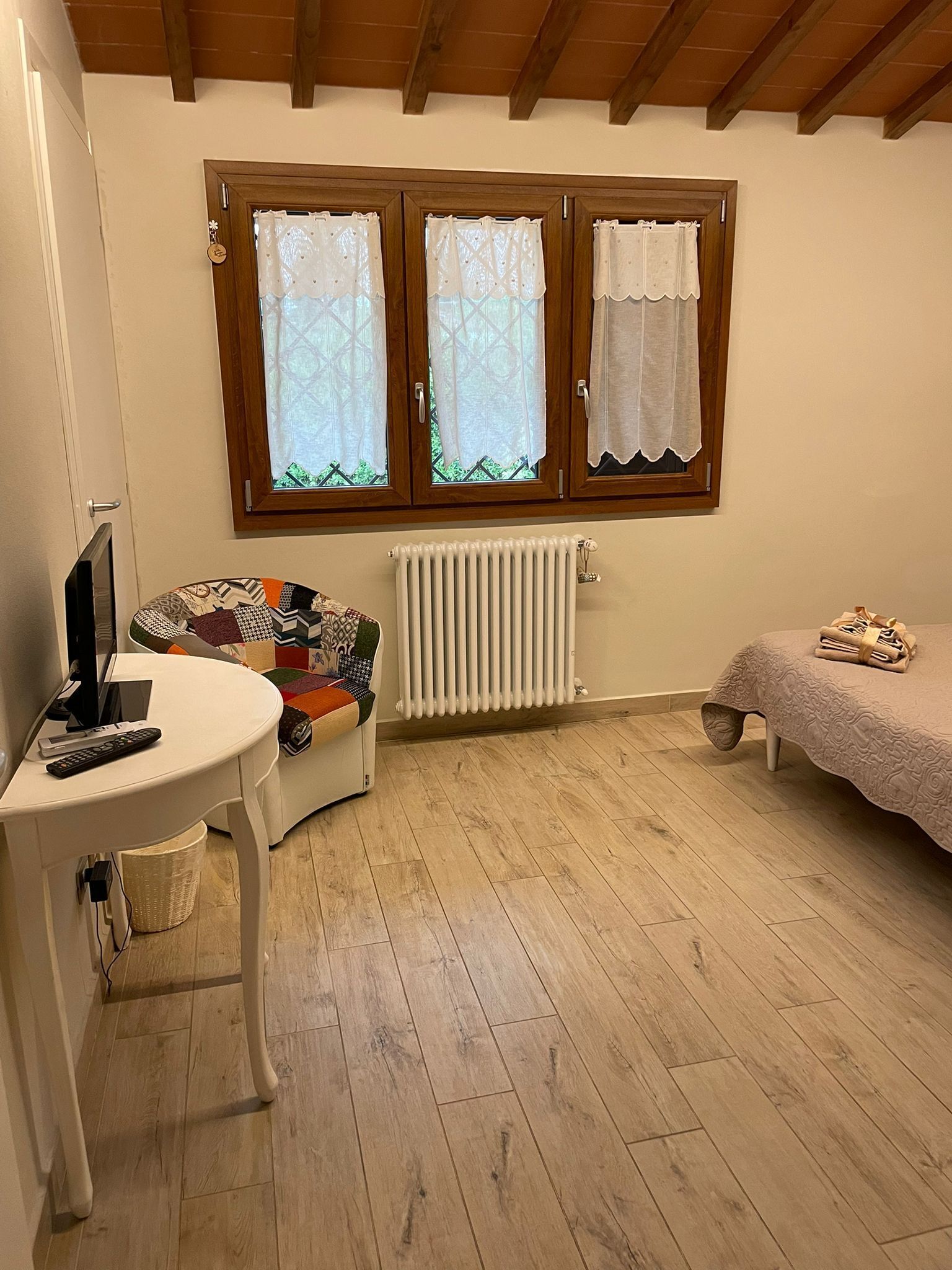 Ein gemütliches Zimmer mit Holzbalkendecke, einem weißen Heizkörper, einem kleinen weißen Tisch mit Fernseher und Fernbedienung, einem farbenfrohen Sofa und einem Bett mit einer beigen Decke.
