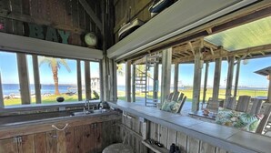 A rustic kitchen with wooden cabinets, a sink, and stools. There is a view of the ocean through large windows. - Beach House on the Bay - Fisherman's Paradise on the bay! (Port Bolivar)