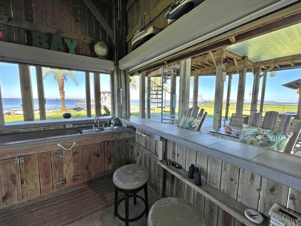A rustic kitchen with wooden cabinets, a sink, and stools. There is a view of the ocean through large windows. - Beach House on the Bay - Fisherman's Paradise on the bay! (Port Bolivar)