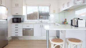 Una cocina moderna con armarios blancos, un refrigerador de acero inoxidable, un microondas, un lavavajillas y una cafetera. Hay una ventana con vistas a un tejado con baldosas.