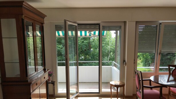 A room with a wooden cabinet, a dining table with chairs, a small stool, and a sliding glass door leading to a balcony with a view of greenery. - Bijou Apartment (Zollikofen)