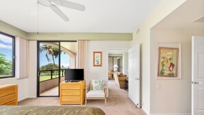 A bedroom with a bed, a TV, a dresser, and a sliding glass door leading to a balcony with a view of greenery. - Summer Deal Golfers Paradise Condo with Fairway and Ocean Views (Waikoloa)
