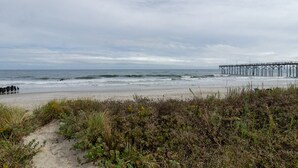 Una playa con un muelle que se adentra en el océano, dunas arenosas y cielo nublado.