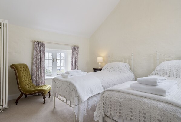 A bedroom with two beds, a green armchair, a window with curtains, and a bedside table with a lamp.