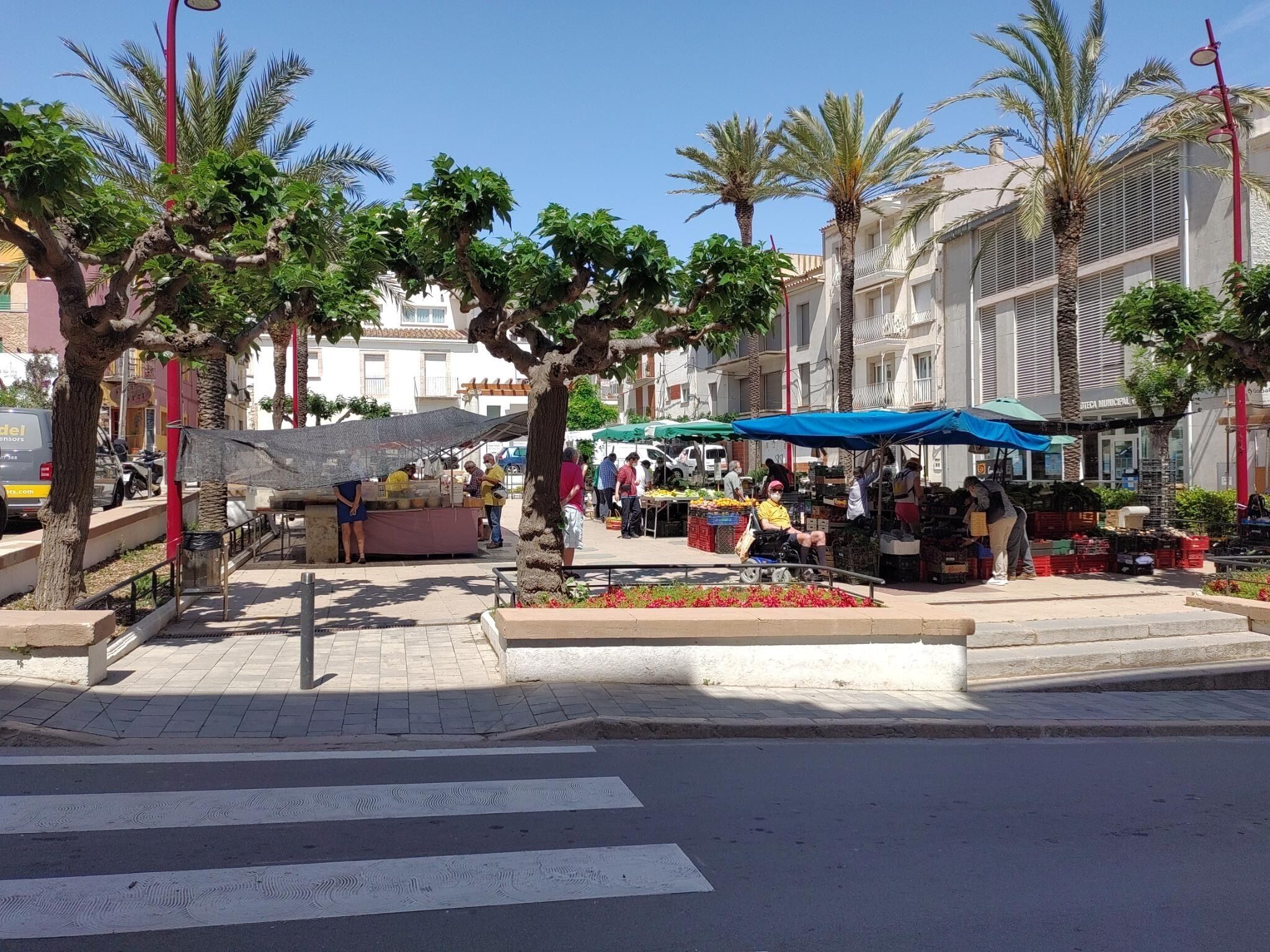 Un mercat de carrer amb venedors i compradors, palmeres i edificis al fons.