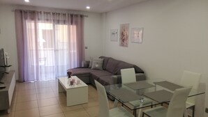 Ein modernes Wohnzimmer mit einem Glastisch, weißen Stühlen, einem grauen Sofa und einem Fernseher auf einem Schrank.