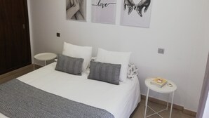 Ein ordentlich bezogenes Bett mit weißer und grauer Bettwäsche, ein kleiner Nachttisch mit einem Buch sowie drei gerahmte Kunstwerke an der Wand.