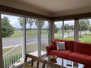 A room with a red sofa, a glass coffee table with a bowl of fruit, and large windows offering a view of a street and greenery.