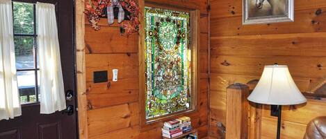 Interior de una cabaña de madera con un ventanal de vitral, una puerta con cortinas, un cuadro enmarcado de un caballo, una lámpara y una pequeña estantería.