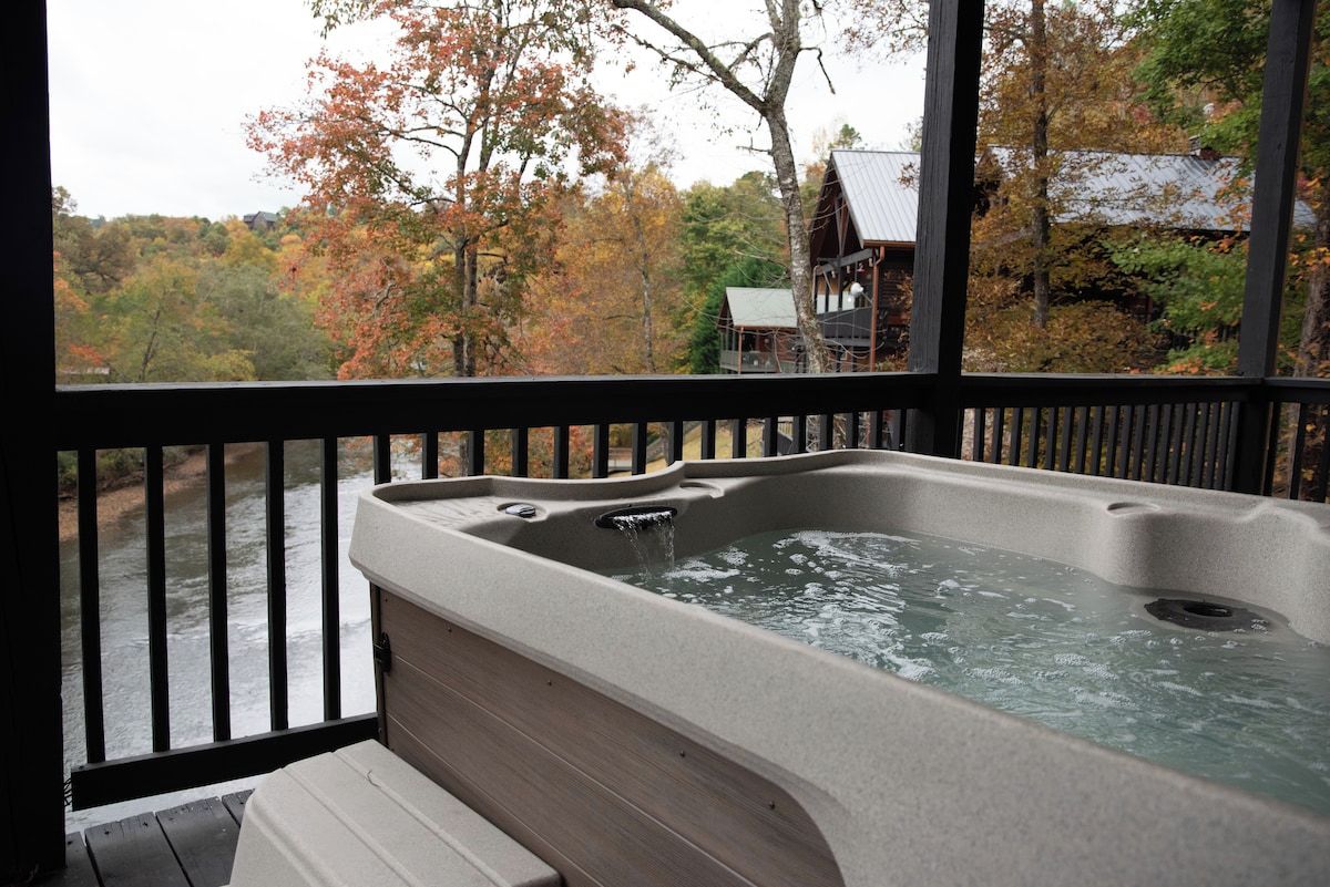 A hot tub on a wooden deck with a view of a river and autumn trees.
