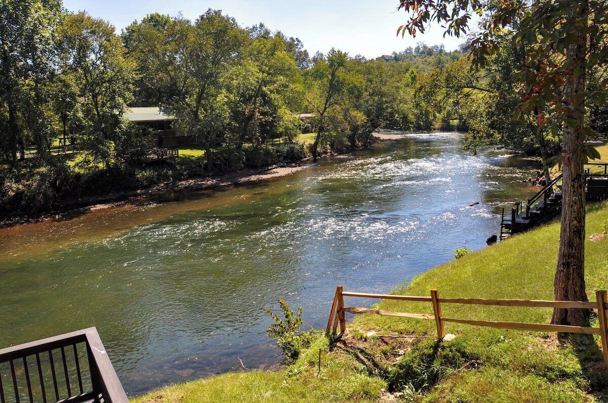 A river with a wooden deck and railing overlooking it, surrounded by trees and a grassy area.