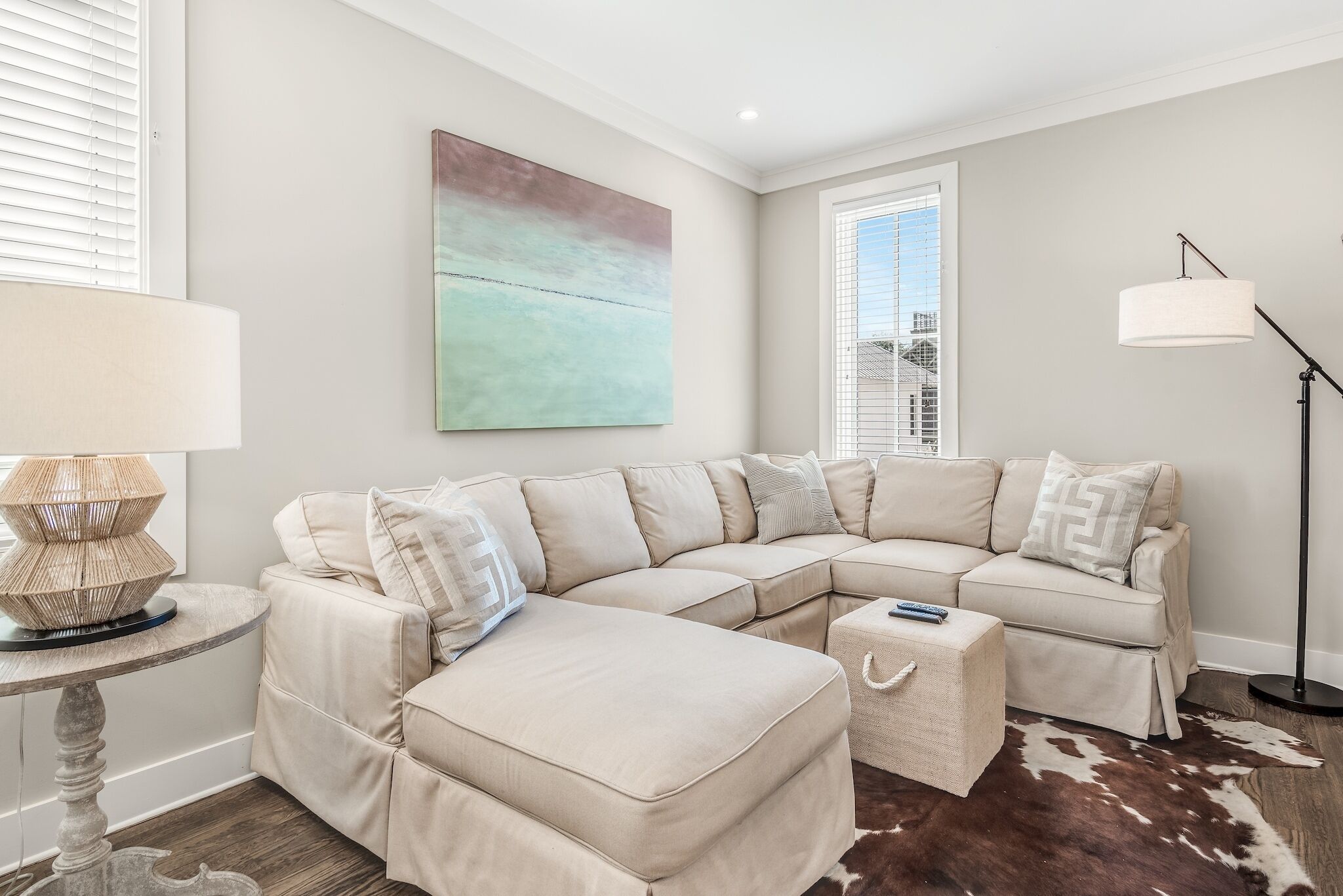 A modern living room with a beige sectional sofa, a round table with a lamp, a cowhide rug, and a large abstract painting on the wall.