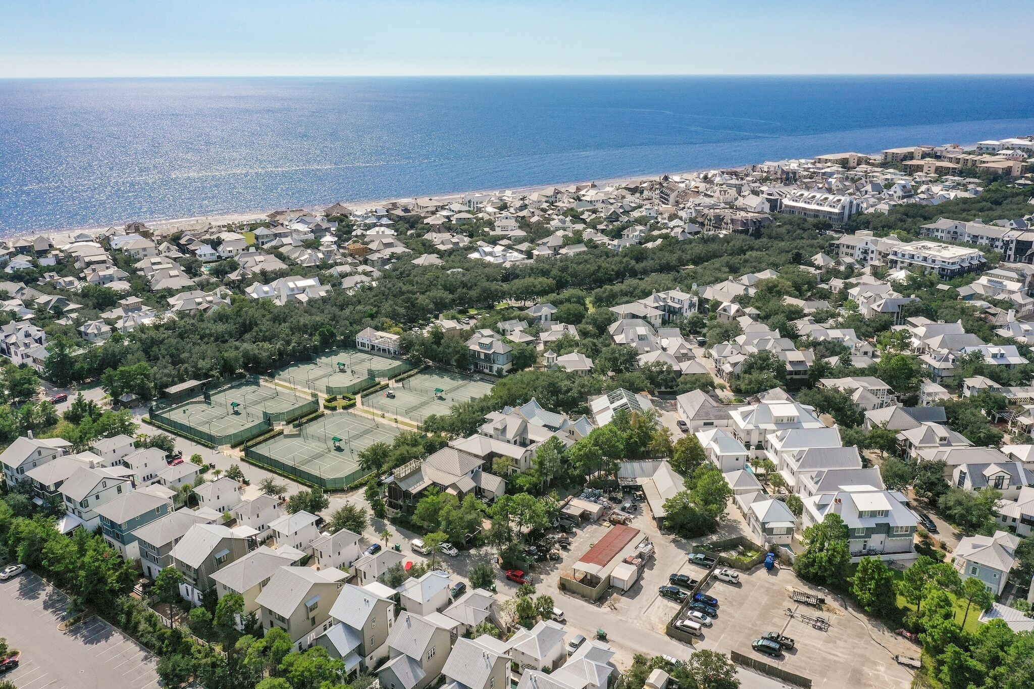 An aerial view of a coastal residential area with houses, a beach, and tennis courts.