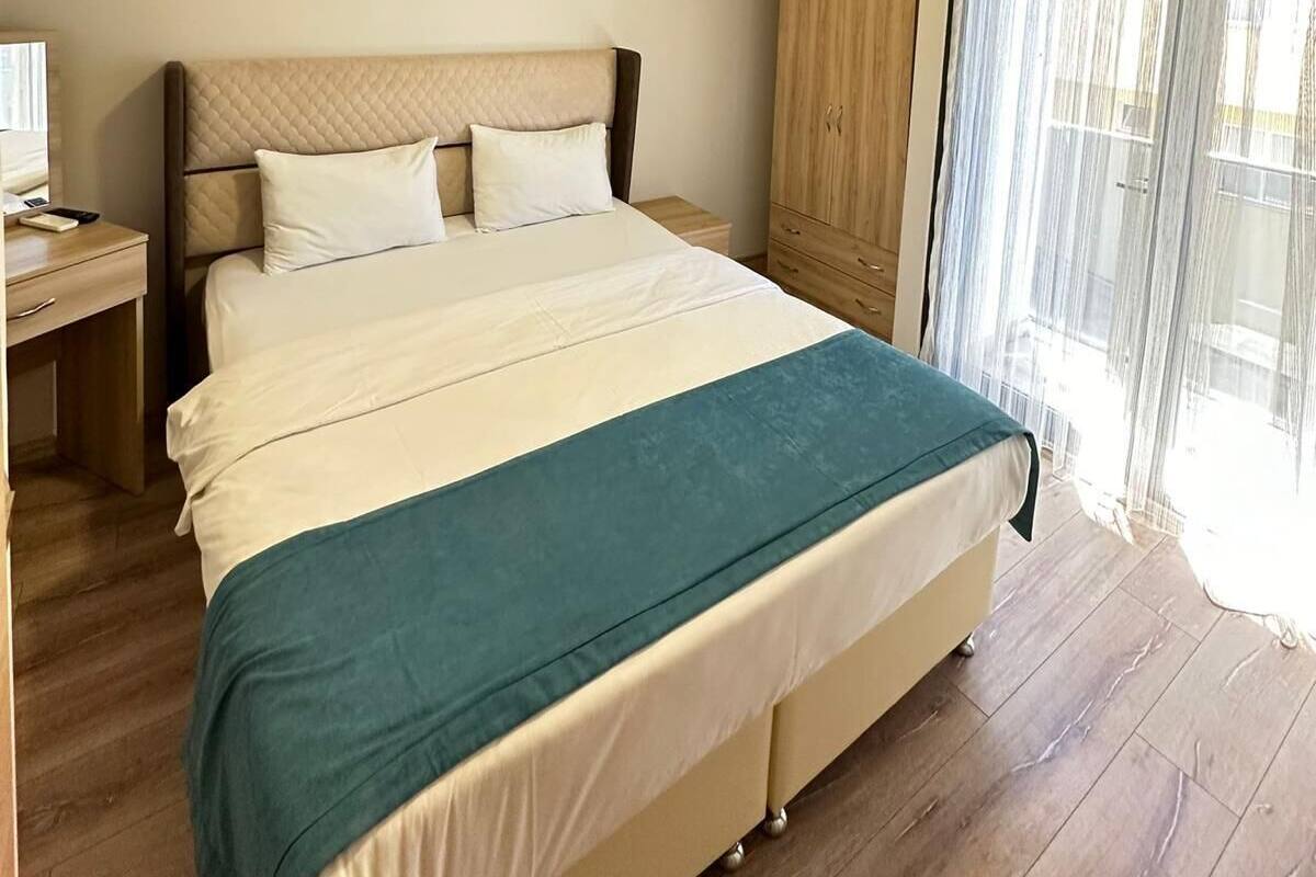 A modern hotel room with a large bed, a desk, a wardrobe, and a window with sheer curtains.