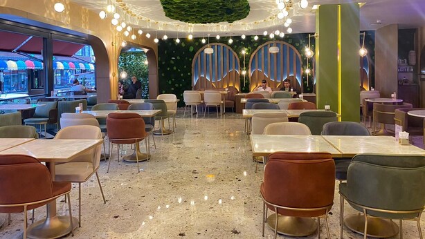 Un restaurante con un interior moderno, que cuenta con mesas y sillas redondas, una pared verde con textura similar al musgo y luces colgantes en el techo.