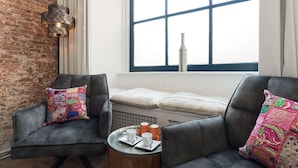 Een kamer met twee grijze fauteuils, een klein rond tafeltje met kopjes en een fles, een bakstenen muur en een groot raam met uitzicht.