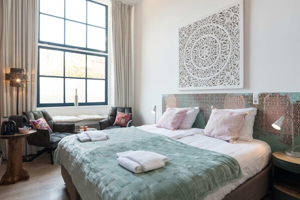 Habitación de hotel con un ventanal grande, una cama con colcha verde, una silla con cojín, una mesita con tetera y una obra de arte en la pared.