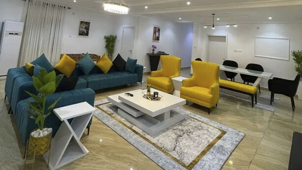 A modern living room with a blue sofa, yellow armchairs, a white coffee table, and a dining area with a white table and black chairs. - Lovely luxury 3 bedroom apartment (Lagos)