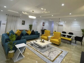 A modern living room with a blue sofa, yellow armchairs, a white coffee table, and a dining area with a white table and black chairs. - Lovely luxury 3 bedroom apartment (Lagos)