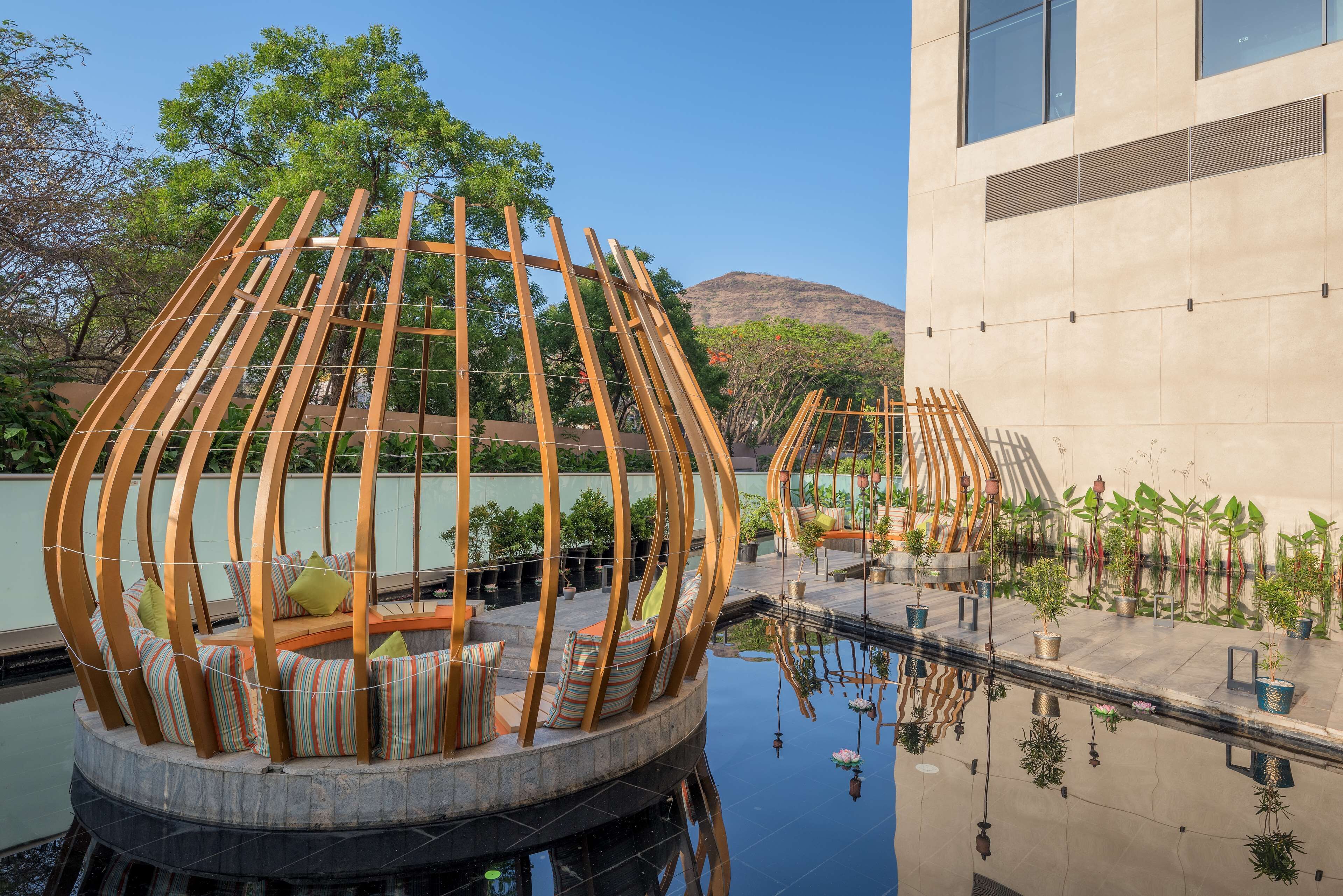 A circular seating area with wooden slats and cushions, surrounded by a pool and potted plants.
