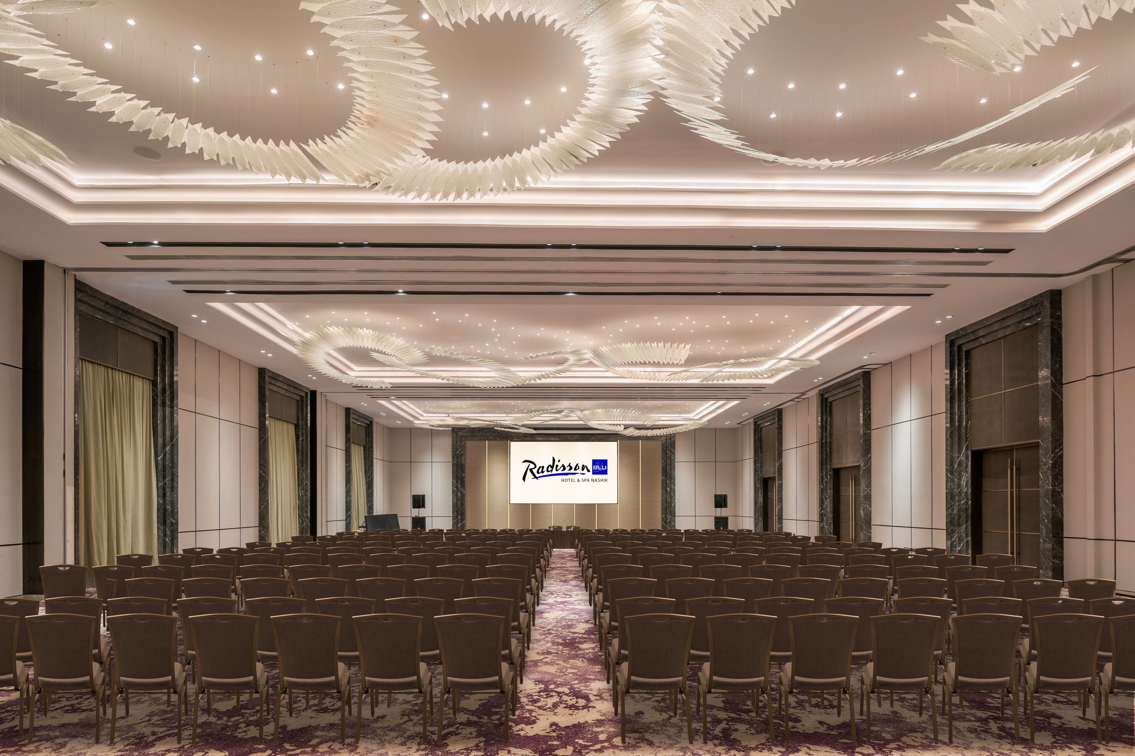A conference room with rows of chairs, a large screen displaying Radisson logo, and a decorative ceiling.