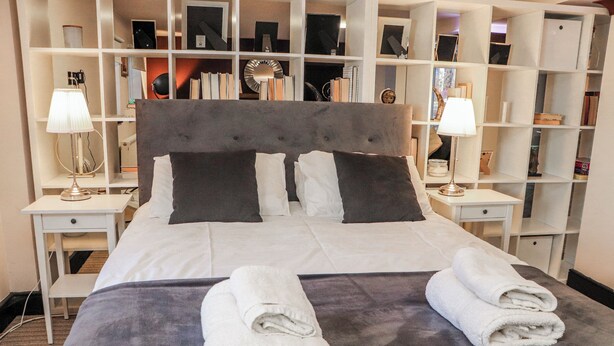 Una cama bien hecha con dos almohadas, una mesita de noche con una lámpara y una amplia estantería al fondo.