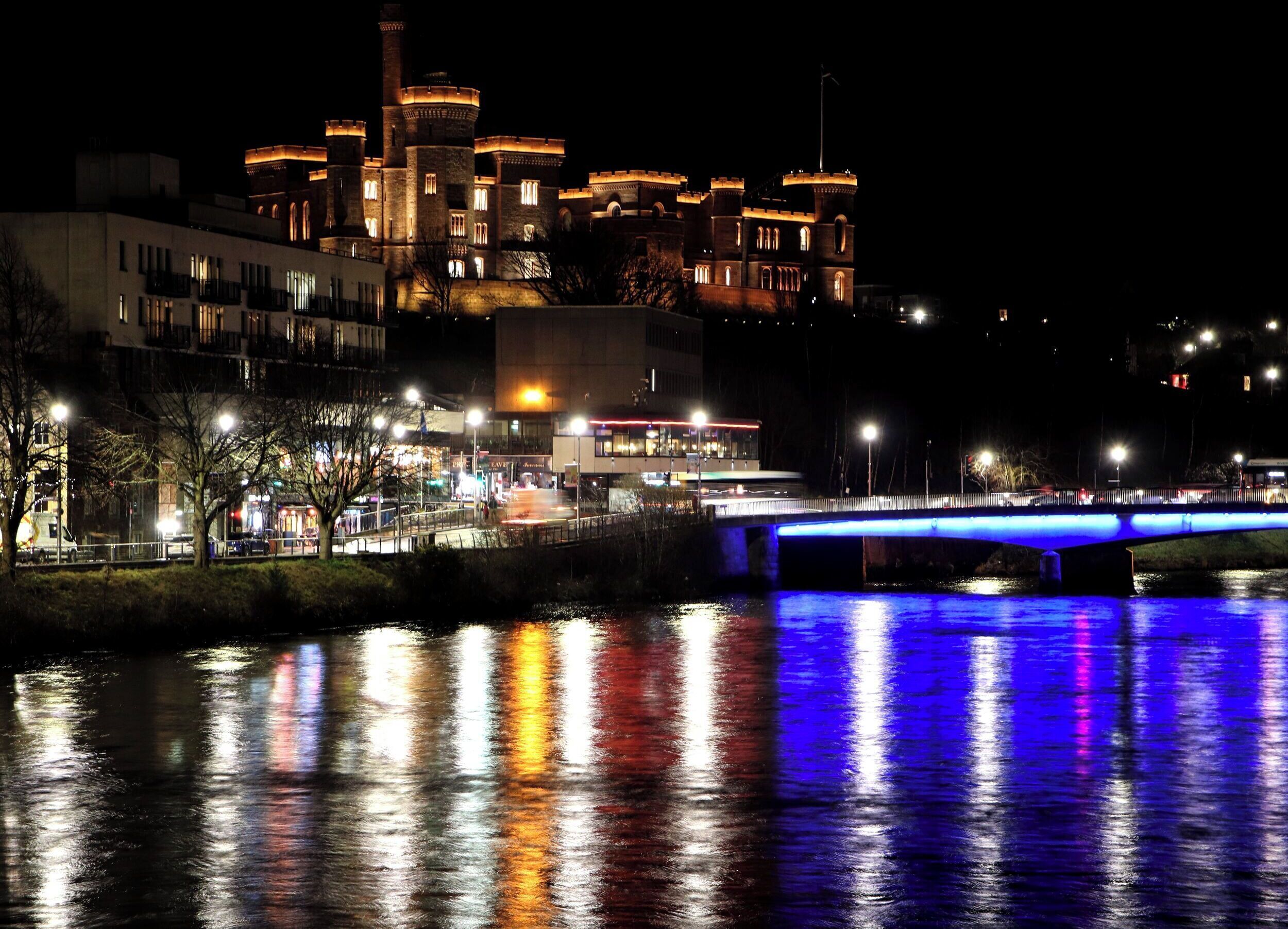 A nighttime view of a castle-like building with a bridge over a reflective water body.