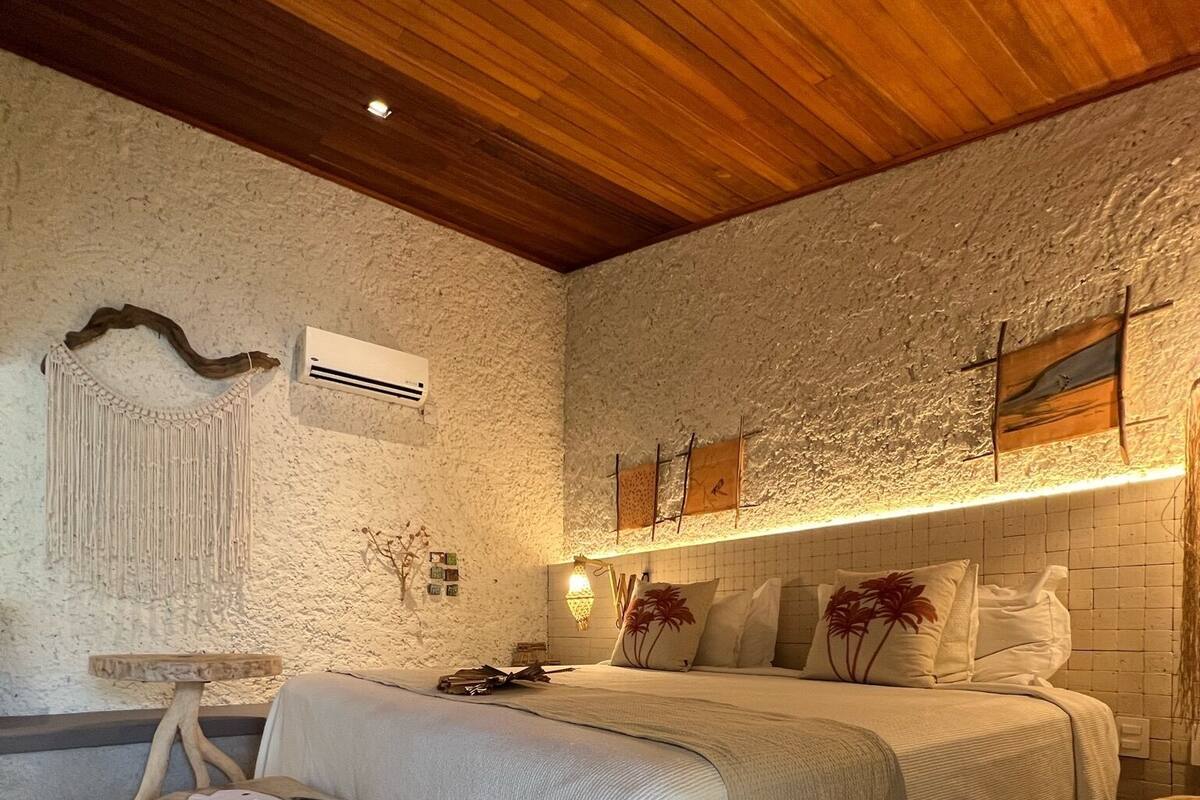 A bedroom with a wooden ceiling, a bed with white bedding, a wall-mounted air conditioner, and a decorative macrame piece.