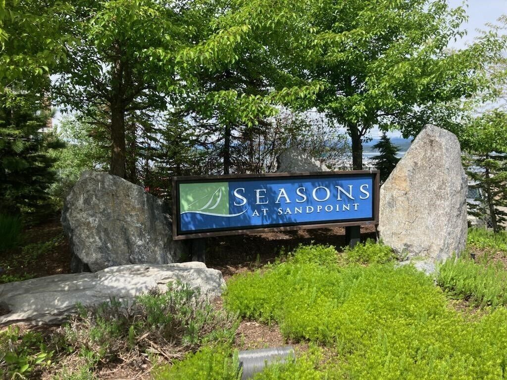 Letrero de 'Seasons at Sandpoint' con un paisaje natural de árboles y rocas.