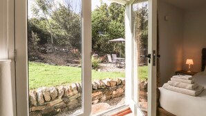 A room with a stone wall, a bed with white linens, and a view of an outdoor garden with a patio and chairs. - Y Felin at Coed Cadw (Crymych)