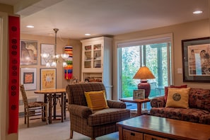 A cozy living room with a plaid armchair, a wooden coffee table, and a sofa. There is a dining table with chairs, a chandelier, and a window with a view of greenery. - Aspen Home - Pet Friendly, Private Hot Tub & Close to Bald Mountain Ski Lodge (Ketchum)