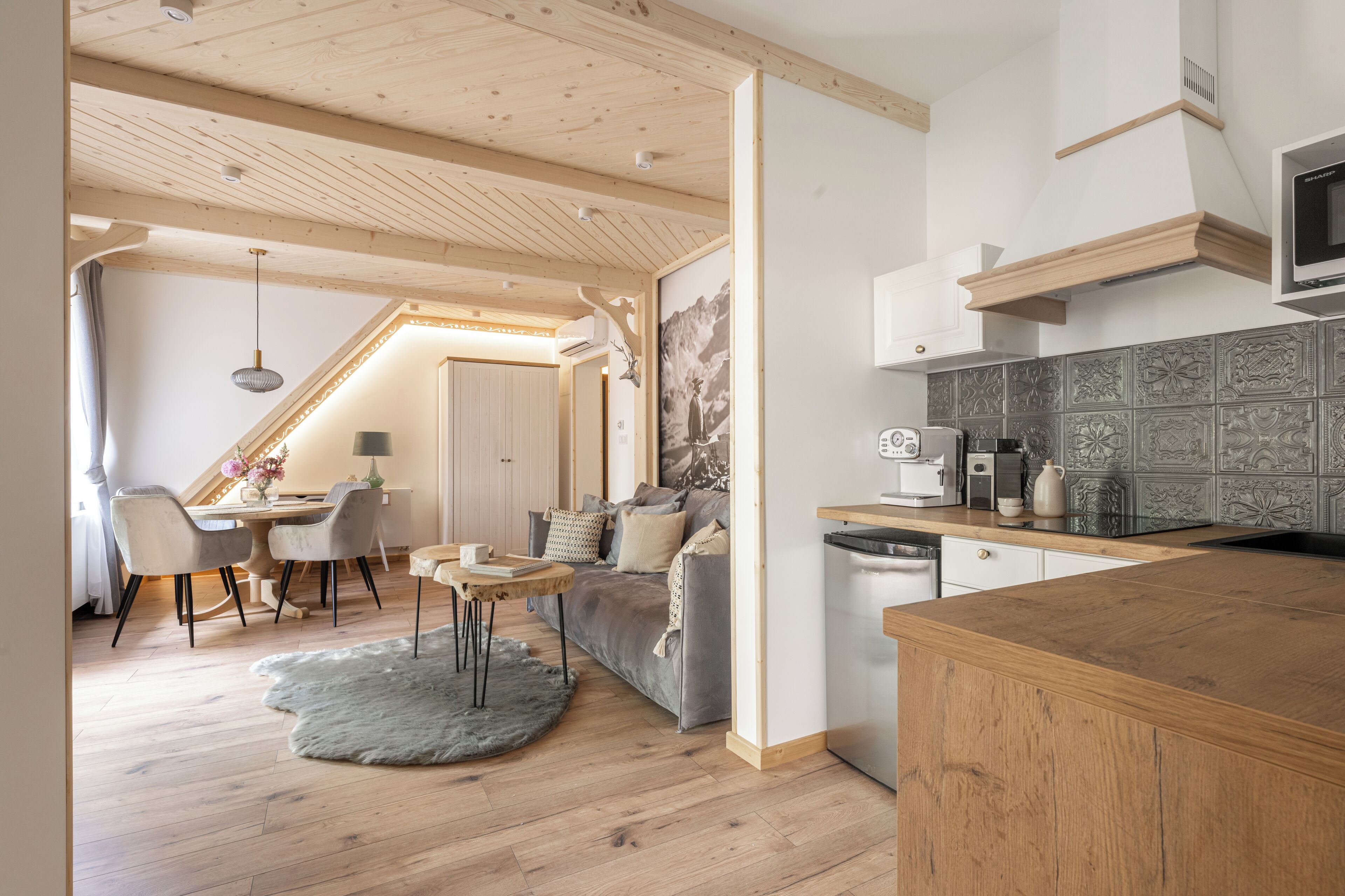 A modern kitchen with wooden cabinets, a built-in oven, and a microwave. There is a dining area with a table and chairs, and a living room with a sofa and a rug.