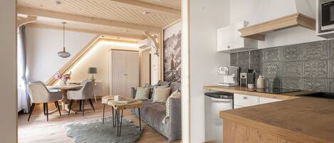 A modern kitchen with wooden cabinets, a built-in oven, and a microwave. There is a dining area with a table and chairs, and a living room with a sofa and a rug.