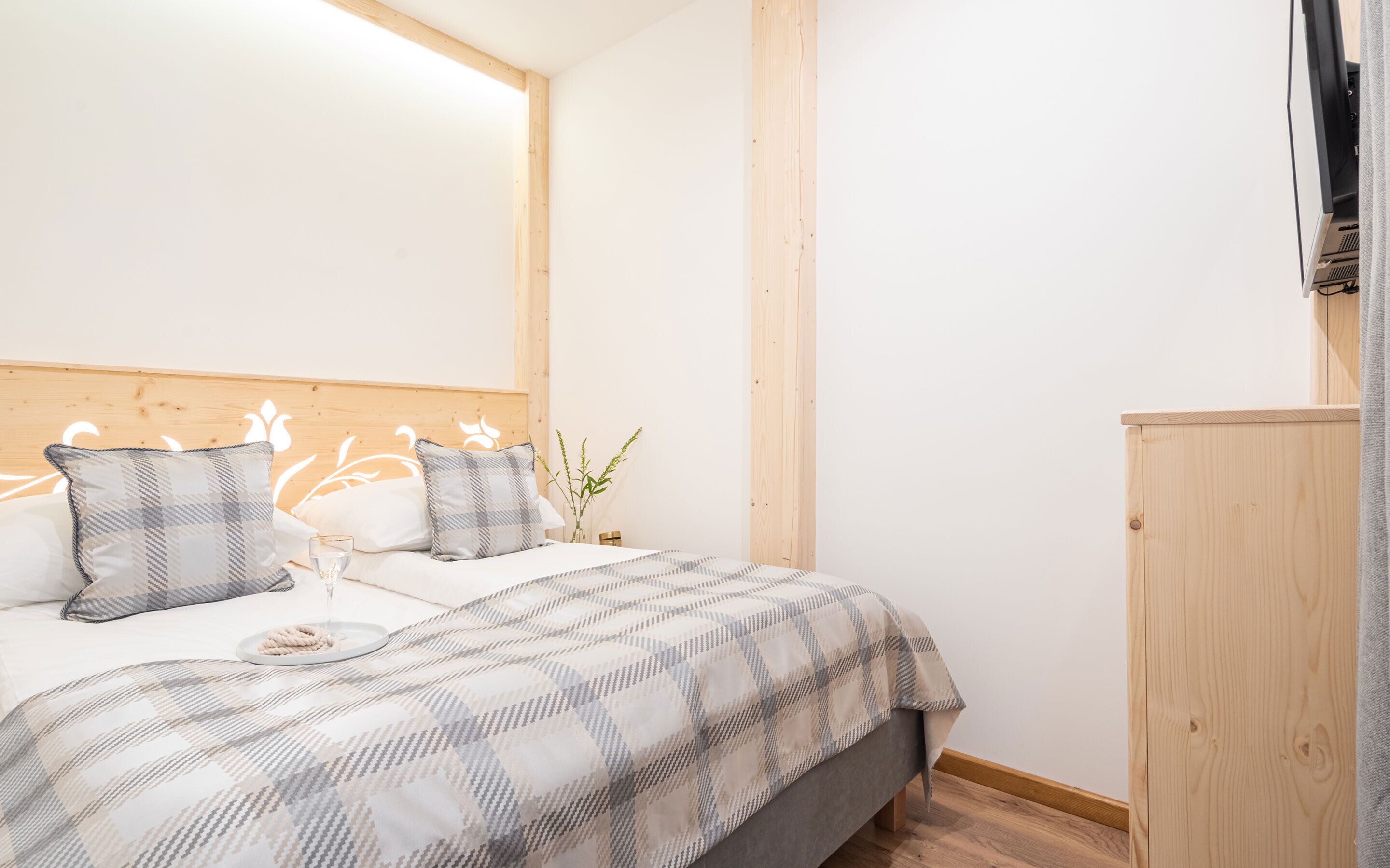 Ein Schlafzimmer mit einem hölzernen Kopfende, einem Bett mit kariertem Überwurf, zwei Kissen, einem Nachttisch mit einem Glas und einer kleinen Pflanze.