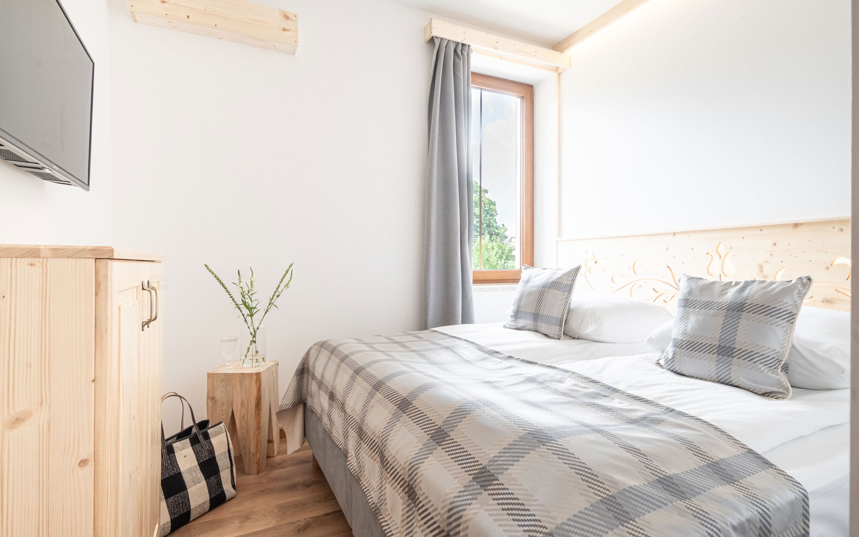 A bedroom with a bed, a wooden wardrobe, a small wooden stool, and a window with curtains.