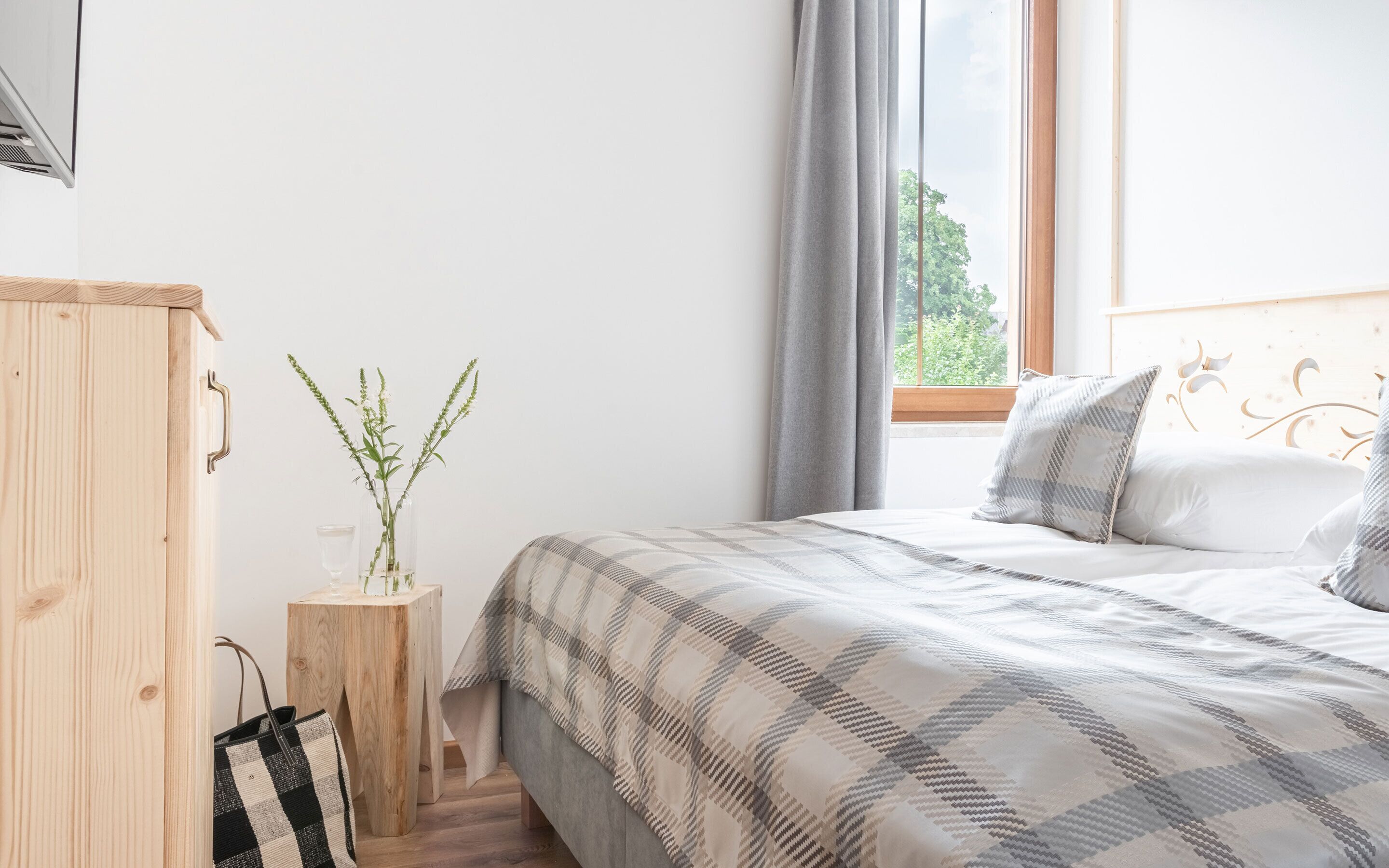 A bedroom with a bed, a wooden wardrobe, a small table with a vase of plants, and a window with curtains.