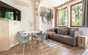 Een gezellige woonkamer met een grijze bank, een ronde tafel met stoelen, en een groot raam met uitzicht op bomen.