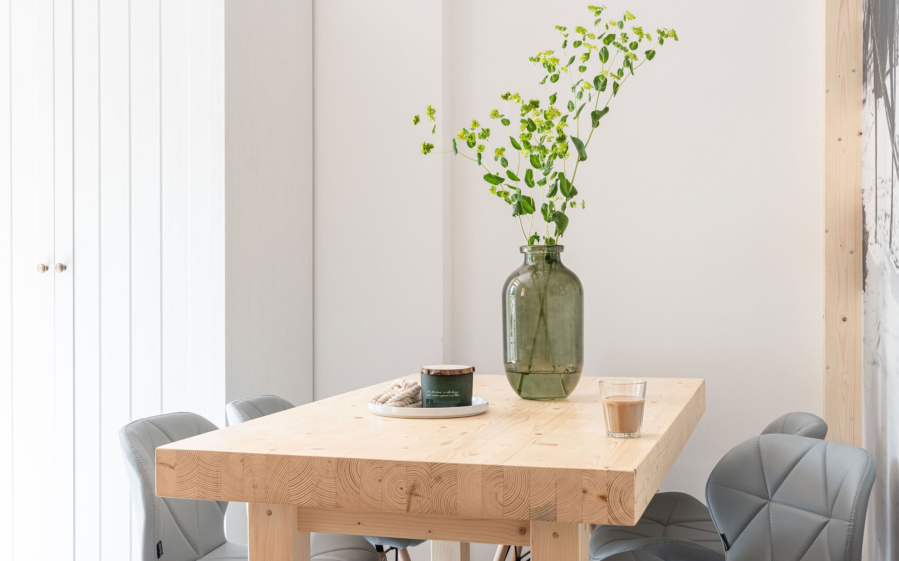 A wooden table with a glass vase containing green plants, a cup, and a glass of a light-colored beverage.